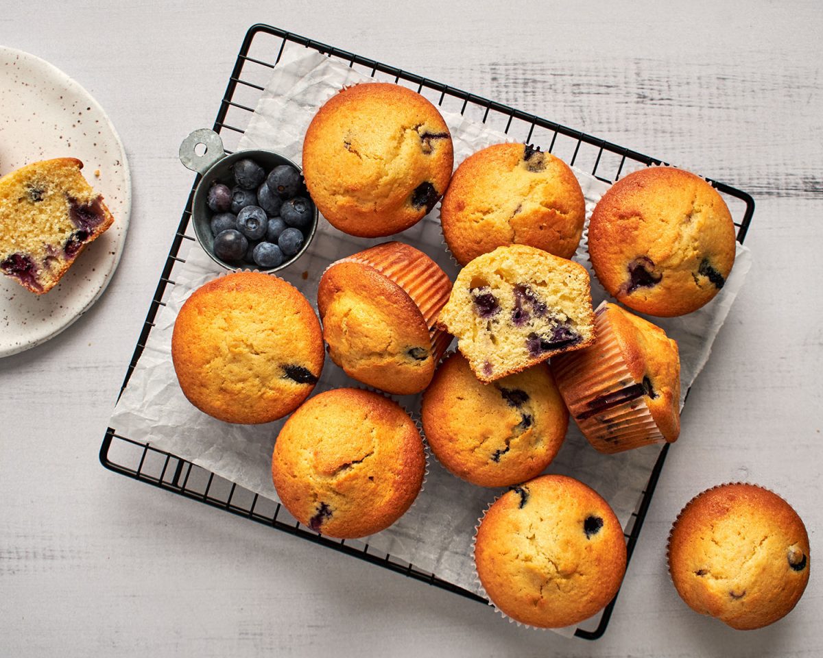 blueberry cornbread muffins piled onto cooking rack with small bowl of blueberries next to the muffins
