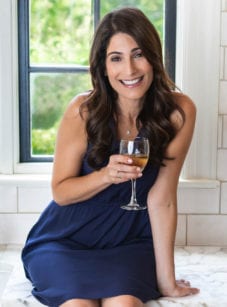 Jenn in a blue dress holding a glass of wine while sitting on a counter.