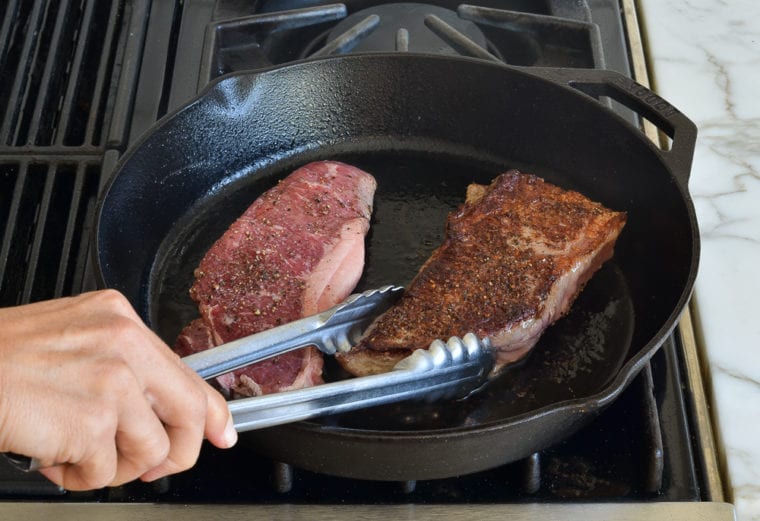 Tongs flipping a steak in a skillet.