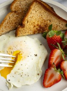 over easy egg, strawberries and toast on white plate with fork in runny yolk