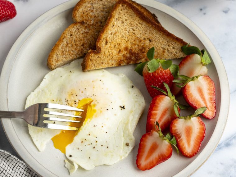 over easy egg, strawberries and toast on white plate with fork in runny yolk