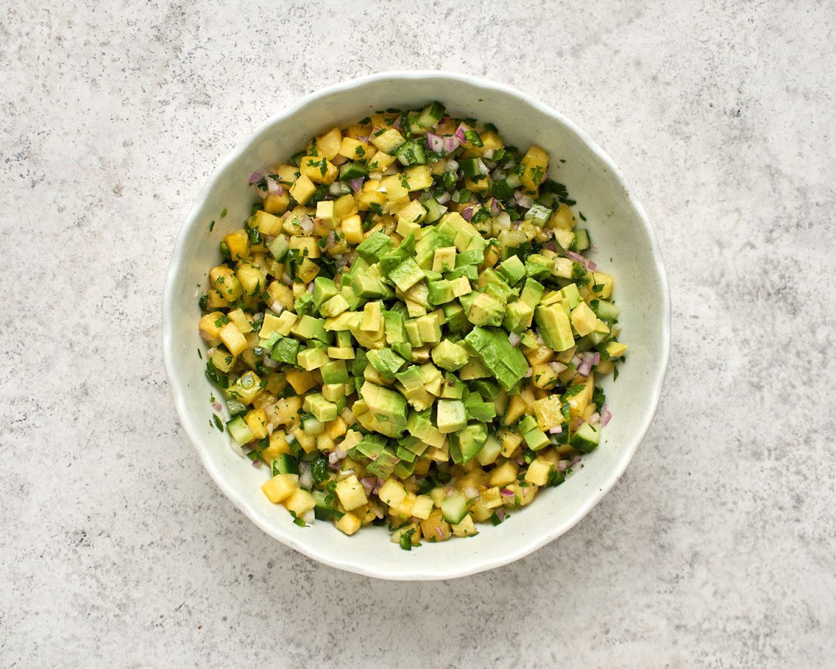 diced avocado on top of combined pineapple, lime juice, onion, cucumber, jalapeño pepper, sugar, vegetable oil, salt, and cilantro in white bowl