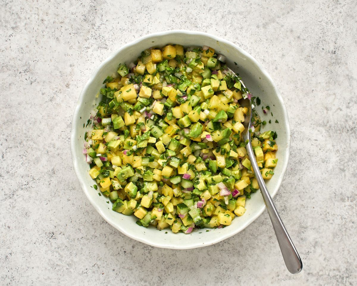 avocado mixed into combined pineapple, lime juice, onion, cucumber, jalapeño pepper, sugar, vegetable oil, salt, and cilantro in white bowl