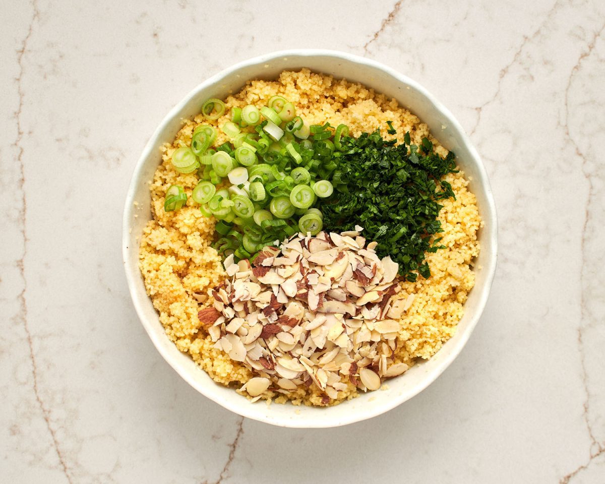 scallions, fresh herbs, and almonds added in piles to cooked couscous in white serving bowl