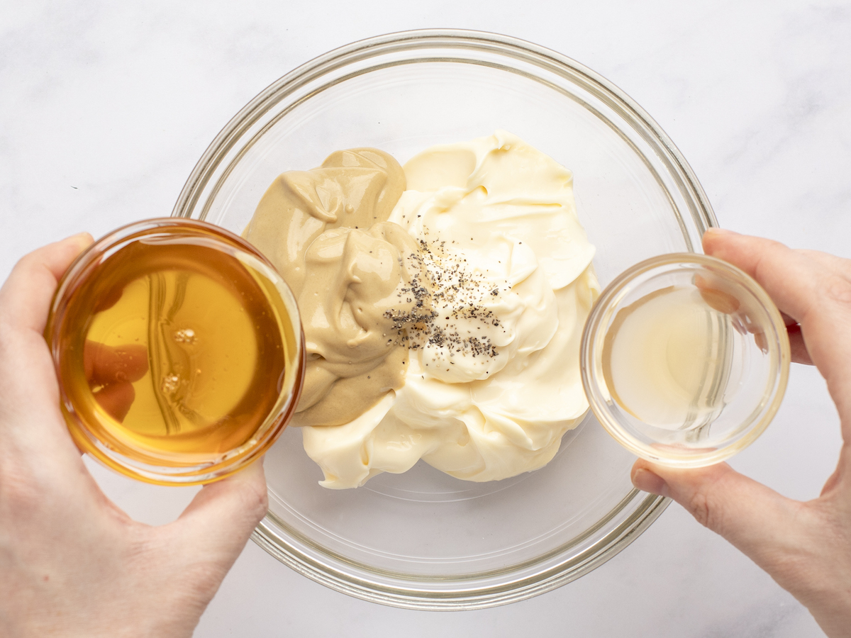 pouring vinegar and honey into glass bowl containing mustard, mayonnaise, and black pepper