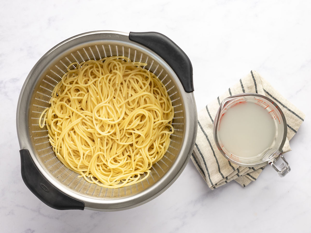 cooked spaghetti in large colander with measuring cup holding pasta cooking liquid to the right of colander