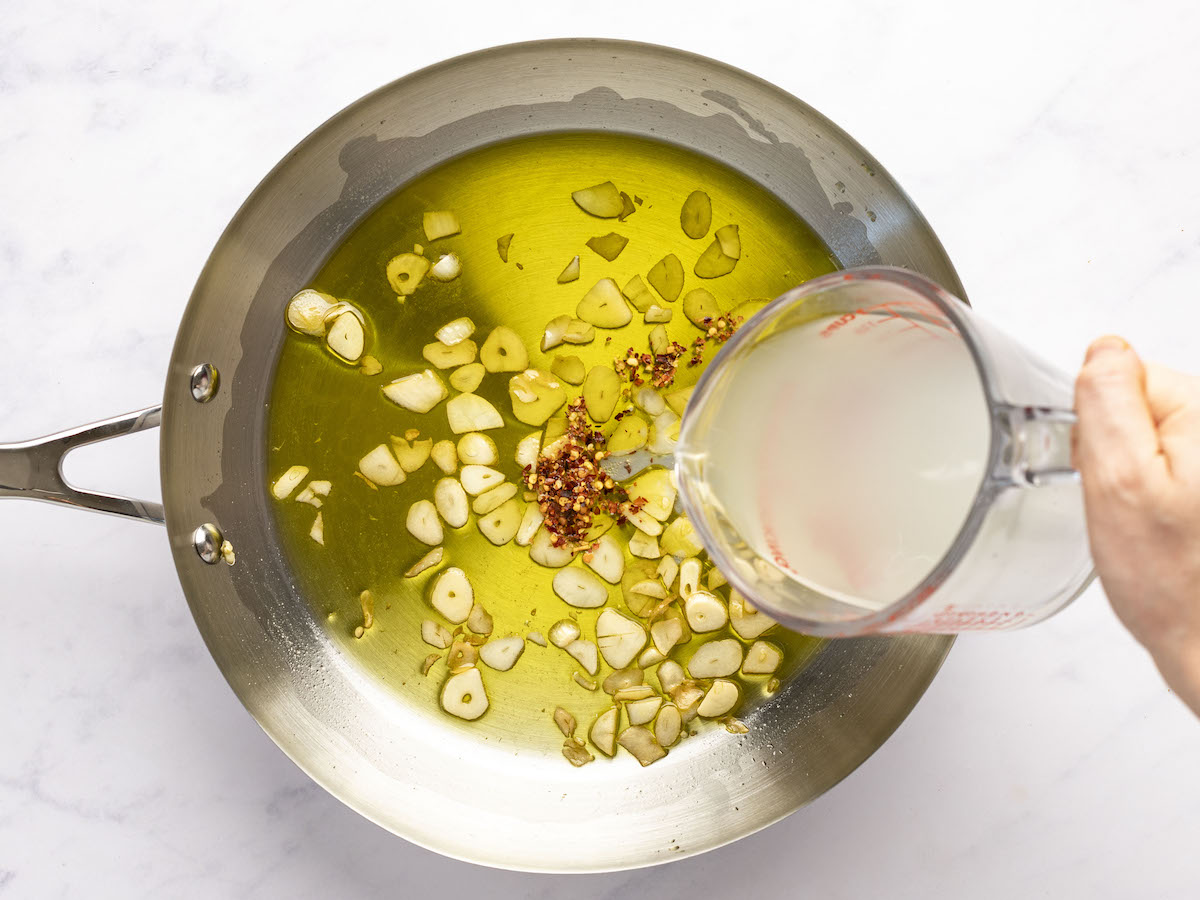 adding pasta cooking liquid into large skillet with olive oil, sliced garlic, and red pepper flakes