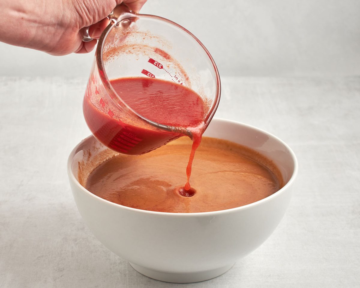 pouring tomato juice into white bowl with finished gazpacho