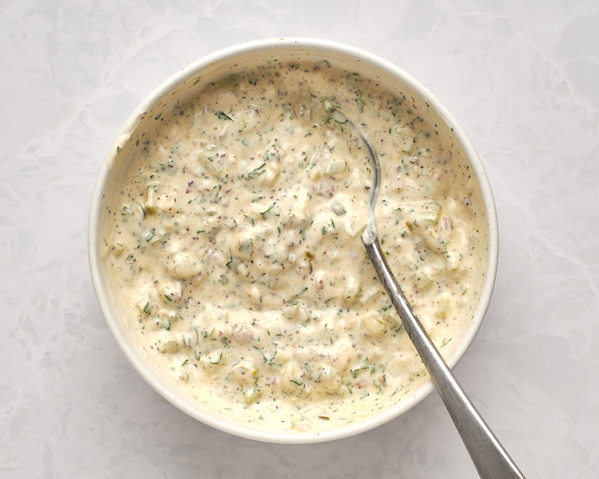 large white bowl with spoon combining celery, onion, relish, mayonnaise, dry mustard, celery seed, parsley, pepper, and salt