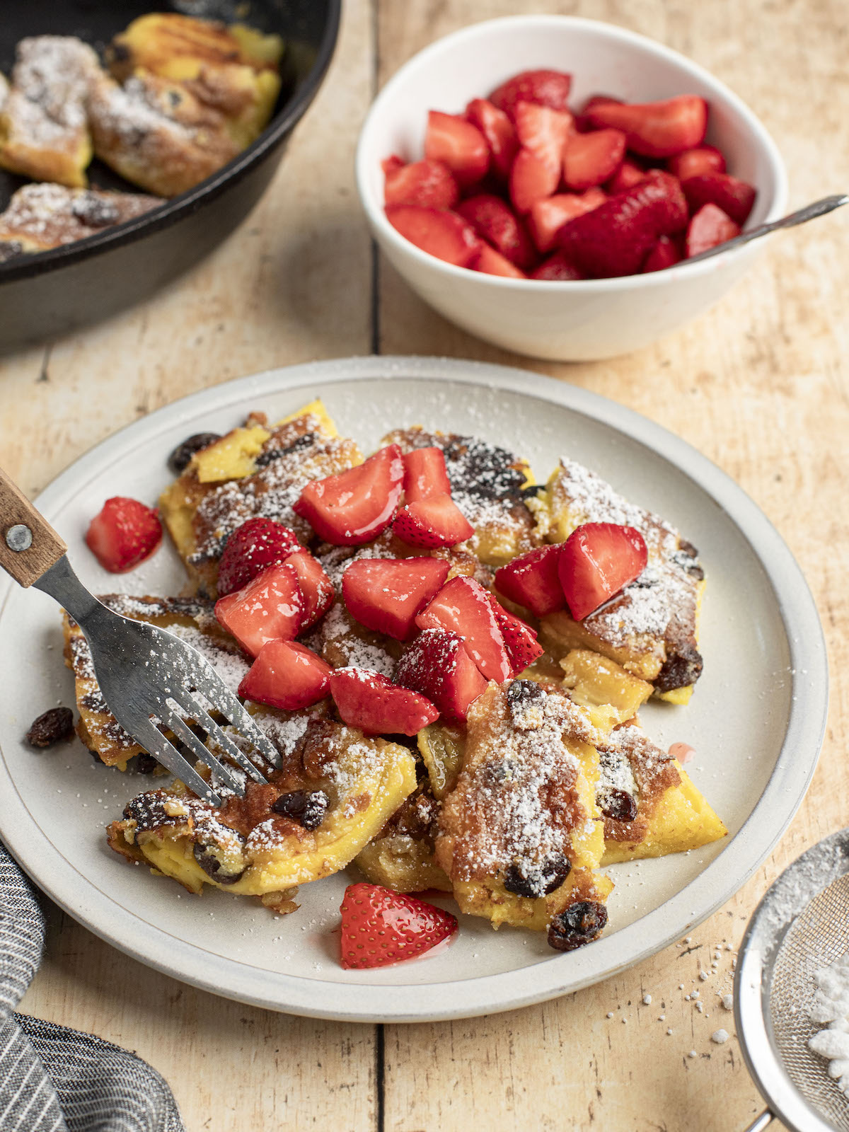 confectioner sugar dusted kaiserschmarrn on white plate with strawberry pieces scattered on top, a white bowl of strawberries and non-stick skillet with remainder of kaiserschmarrn adjacent to the plate 