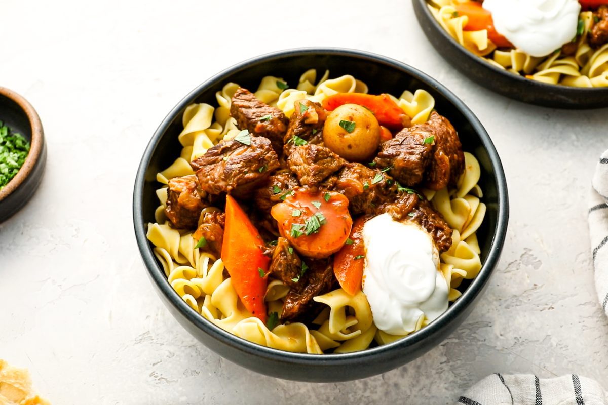 hungarian goulash on top of thick noodles with a spoonful of sour cream on side of bowl