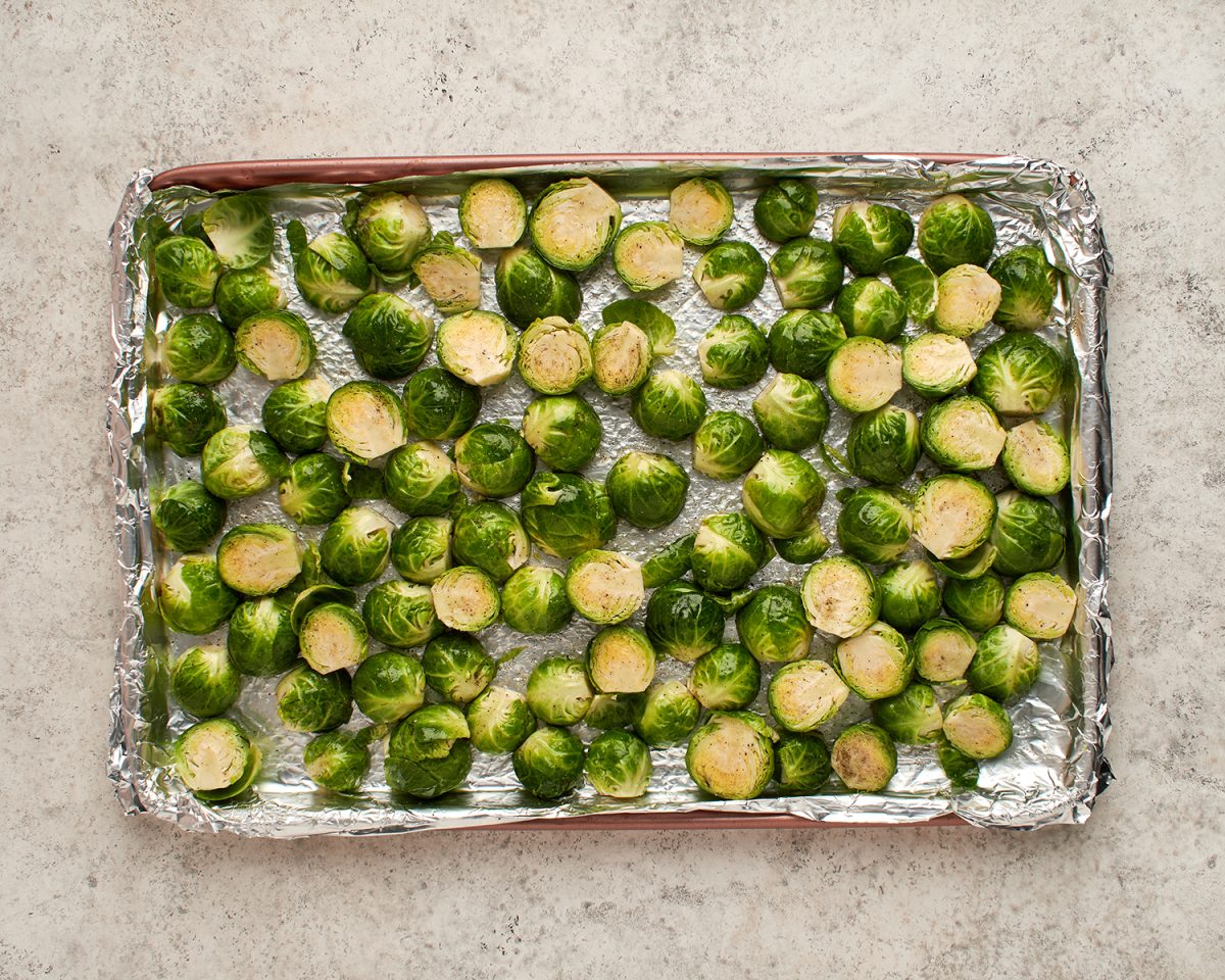 Brussels sprouts tossed with oil, bacon fat, and seasoning on foil-lined baking sheet